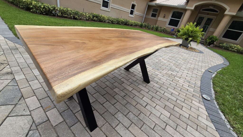 A stunning 6ft live edge dining table crafted from a single 3-inch thick piece of acacia hardwood, featuring natural wood grain and a clear coat sealer. Supported by heavy-duty black steel bases, placed on a brick patio in front of a modern home with green landscaping.